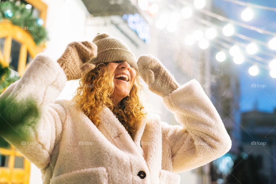 Portrait of young happy woman with curly hair in fur jacket having fun in winter street decorated with lights