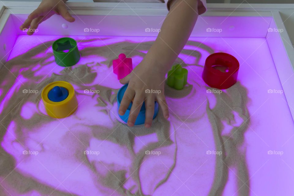 A child plays with colorful toys and sand on a purple backlit table.