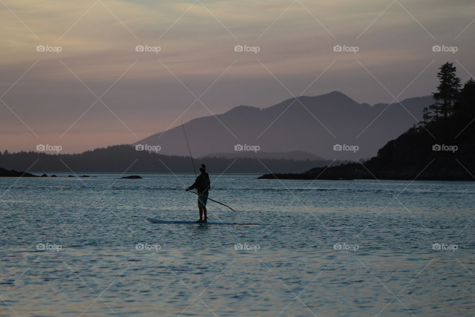 The sun has set behind the mountains & a woman paddling out in the cove to catch a fish or two can be seen silhouetted against the inky blue, calm water. The dark rocky outcrops & purple mountains are silhouetted against the pink & purple sky.