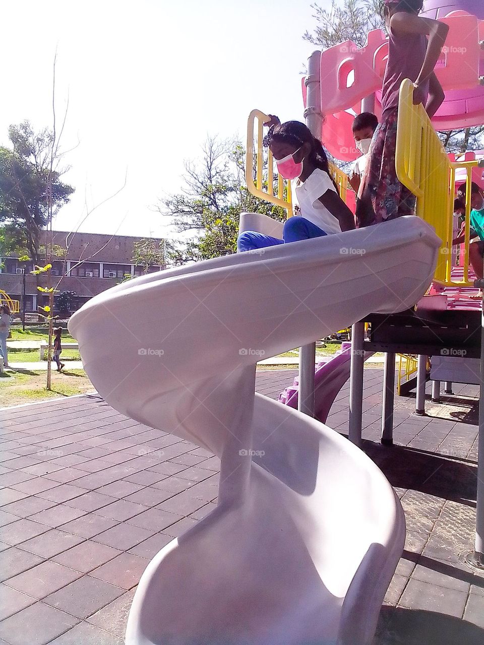 This is a capture of a children's park and children are playing. There is a white colour  curved board and one girl is coming through it. She is wearing a pink colour mask.