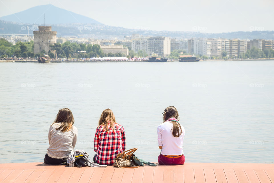 Three Teenagers Girls Friends Sitting On The Wooden Dock And Enjoying The City View In Front Of The Sea
