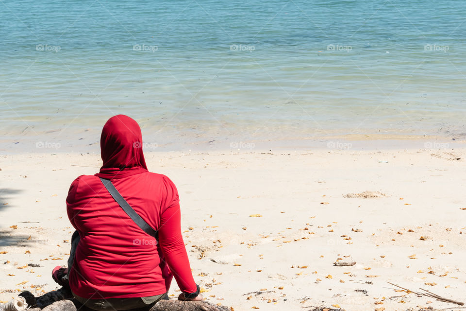 A girl in red, sitting down on the beach in daytime, enjoying the view of nature