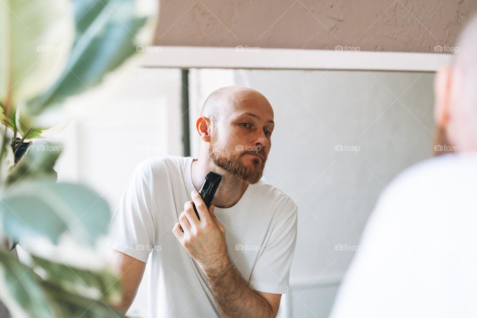 Handsome young bearded man trimming his beard with machine in bathroom at home