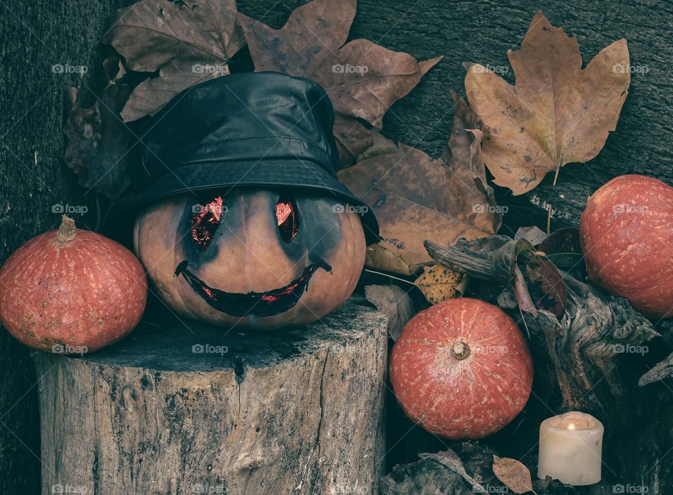 A carved pumpkin with dark glowing, red eyes and wearing a hat, sits on a log surrounded by smaller pumpkins and autumn leaves