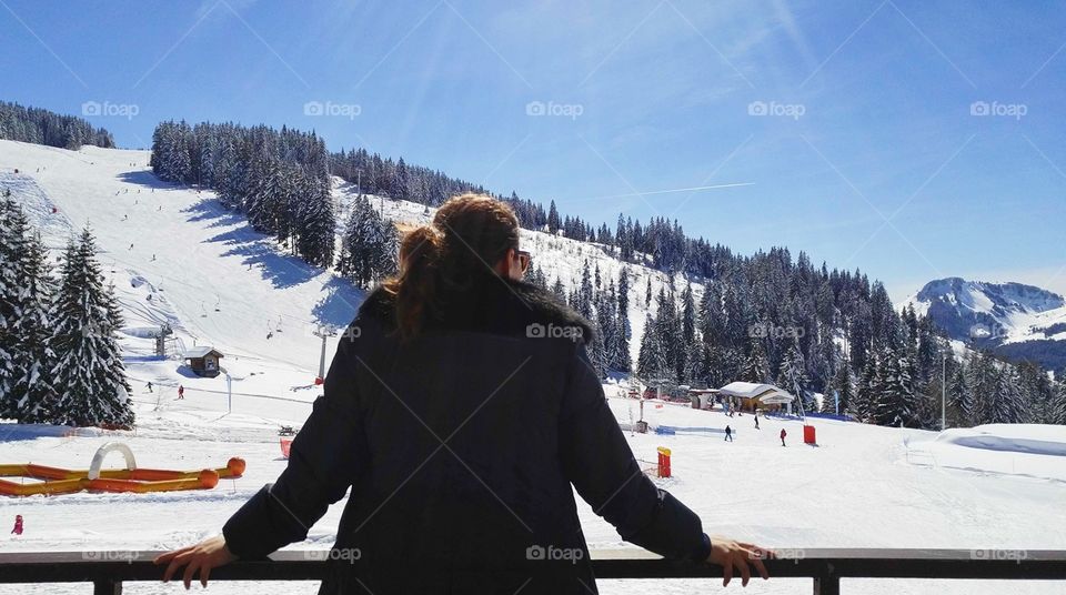 Young woman leaning on a wooden railing and looking out toward a snow-covered ski resort with snow-laden fir trees and snow-capped mountains in the background and the sun's rays shining overhead.