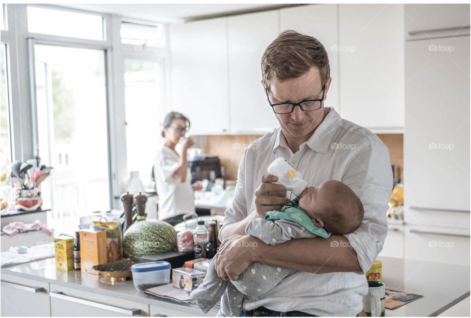 A father feeding baby while mother is in the background 