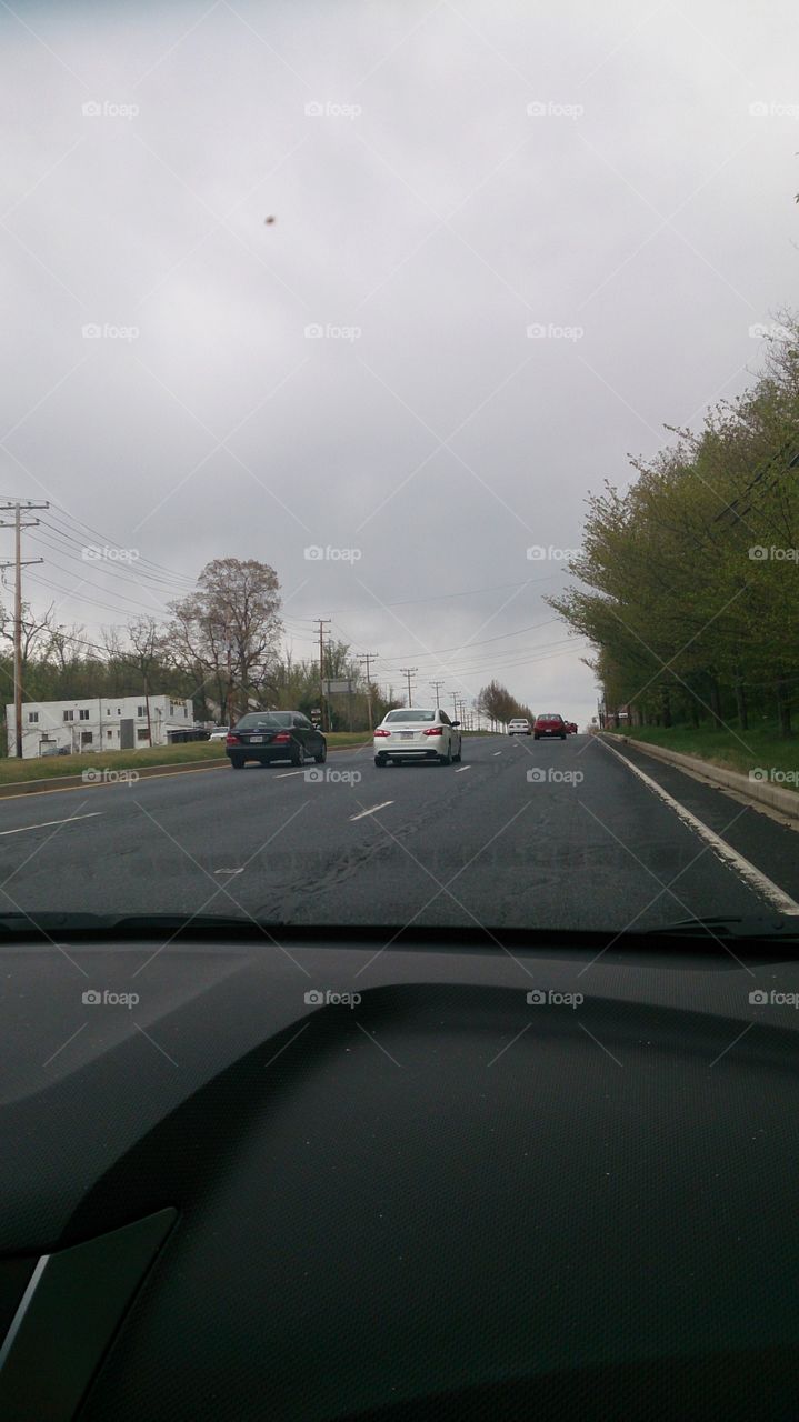 Cars driving down a road during a cloudy day. There's some trees and few cars on the road.