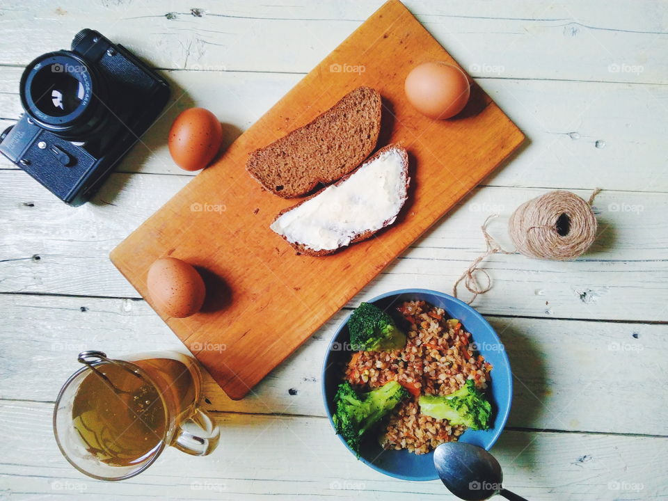 a bowl of buckwheat porridge, bread and butter, eggs, a cup of tea, garlic and a camera on the table