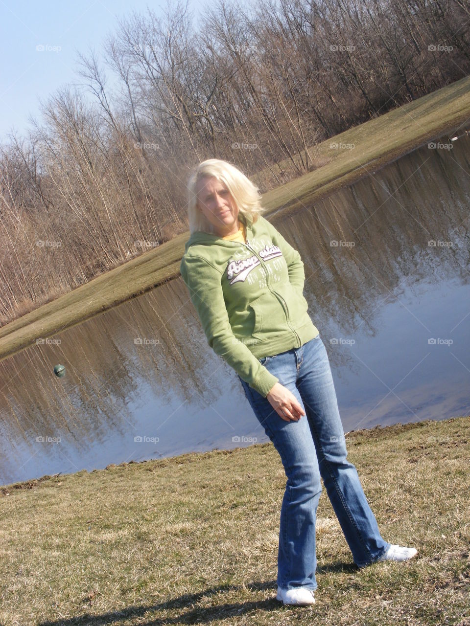Skinny blond woman looking at camera and standing next to a pond.
