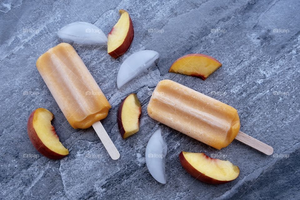 Looking down on a gray marble table top with melting peach juice bars surrounded with ice cubes and slices of peaches.