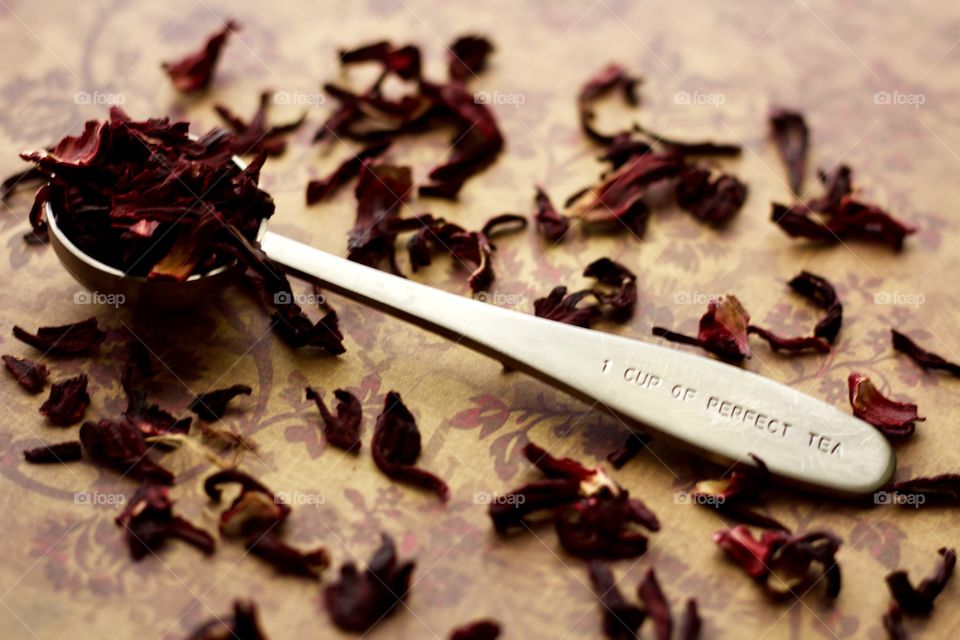 Angled view of a stainless steel measuring spoon imprinted with the phrase, “1 CUP OF PERFECT TEA,” filled with dried hibiscus petals on a brown and burgundy patterned surface with scattered hibiscus tea 