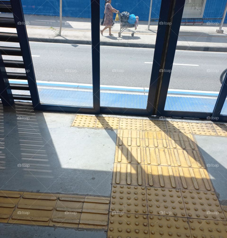 A visão  da janela. Uma senhora, caminha, lentamente, pela calçada, empurrando um carinho de fazer compras dentro do supermecado.