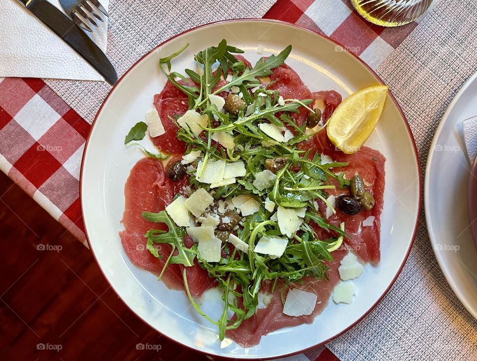 Beef carpaccio in the white plate with lemon and greenery, top view 