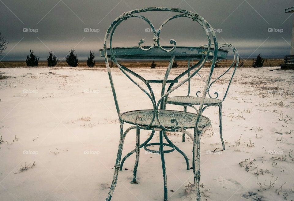 An ice cream table with two chairs sits empty in the snow in winter in an austere background in grays and blues "Table for two"