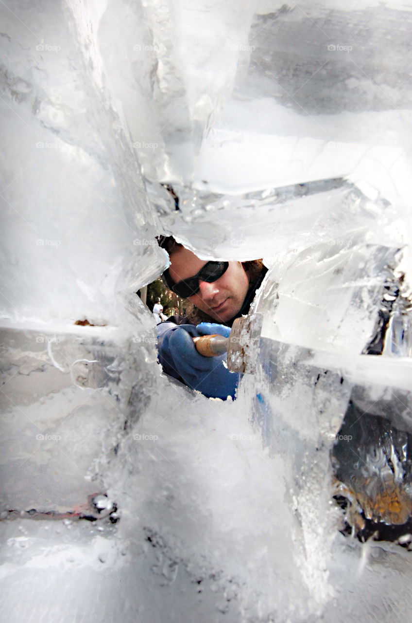 AN ICE SCULPTER WEARING SUNGLASSES IS FRAMED IN A HOLE IN SOLID ICE.
