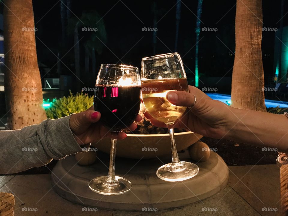 Friends cheers with red wine and white wine in front of an outdoor fire pit and in between two palm trees. 