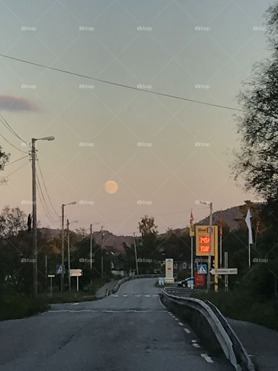 Full moon over a gas station in Norway. The dawn is setting, and the sunset is about to end, as we drive down a road towards the moon, with a gas station to the right. 