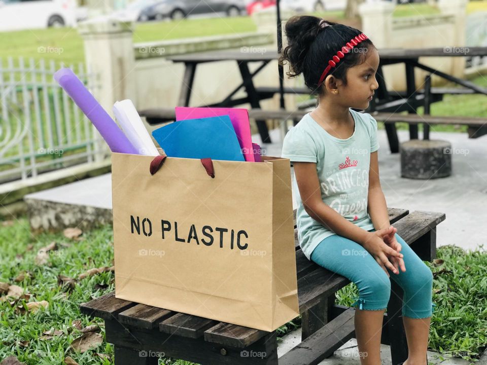 There is a paper bag which wrote NO PLASTIC on its top and the bag contains colour papers and a four year old girl sat beside the bag looking a side.using paper bags reduce the productivity of plastic.