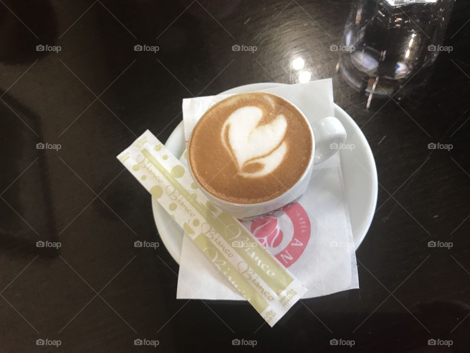 Lovely porcelain cup of coffee with ness coffee in it and with white heart on the top, in the beautiful morning, surrounded by little bags of shugar on a plate.