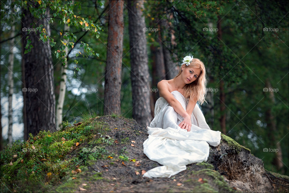 Young beautiful woman in long white dress and white flower in her hair sits on a hill edge of the forest.