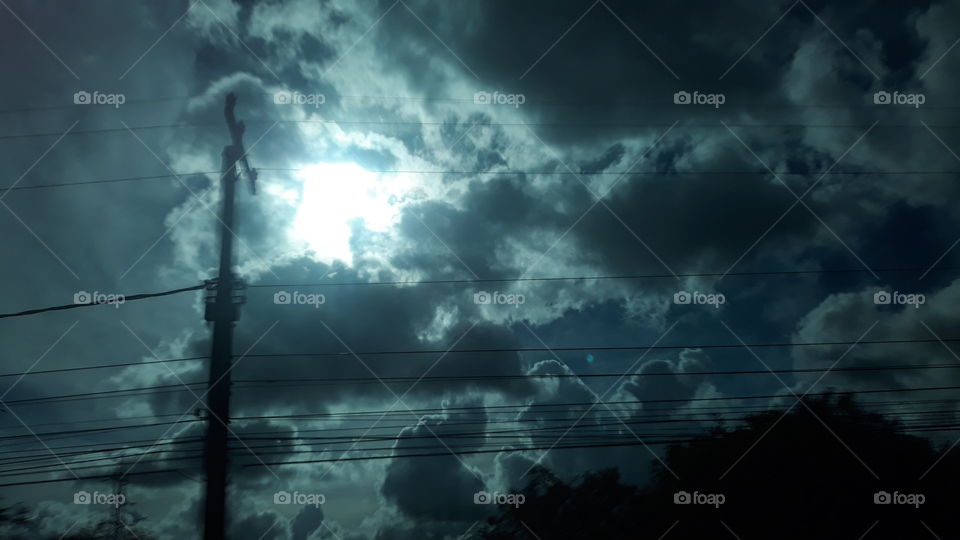 Poste e fios elétricos com o sol, entre nuvens, ai fundo.