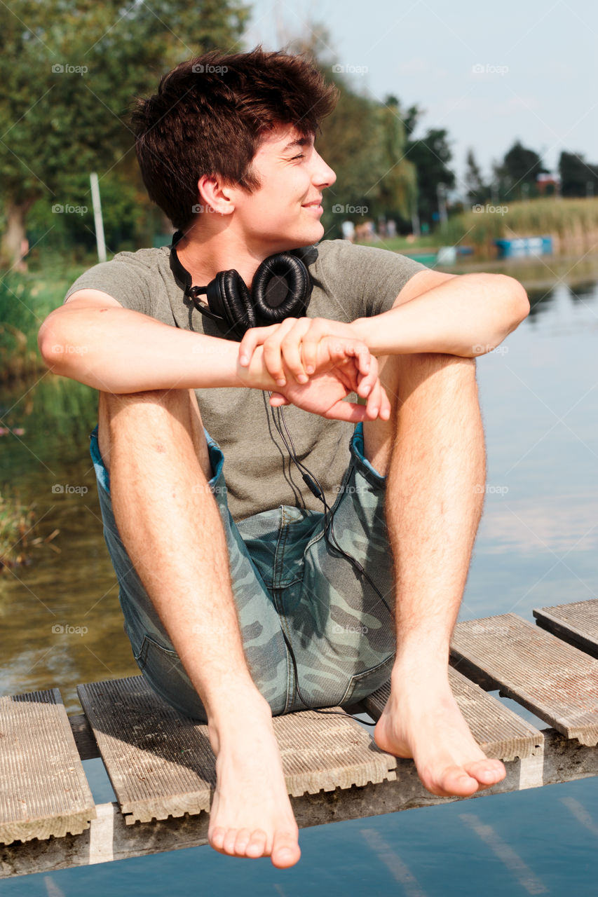 Young smiling boy sitting on jetty over the lake on sunny day in the summertime