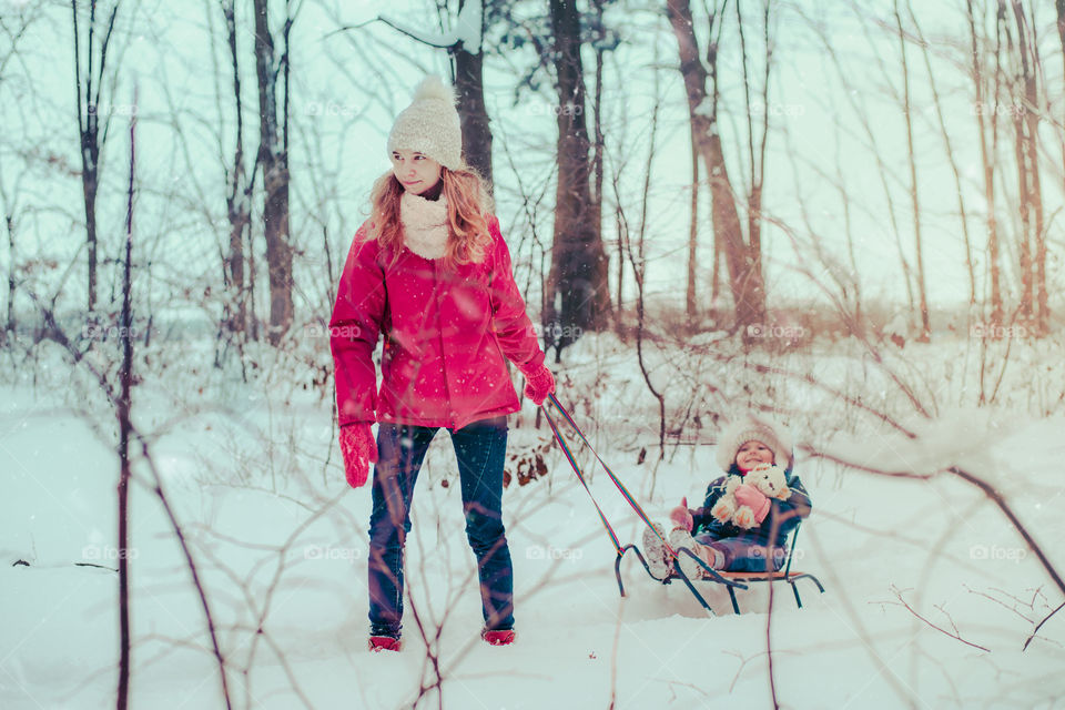 Teenage girl pulling sled with her little sister, a few years old girl, through forest covered by snow while snow falling, enjoying wintertime, spending time together. Girls are wearing winter clothes