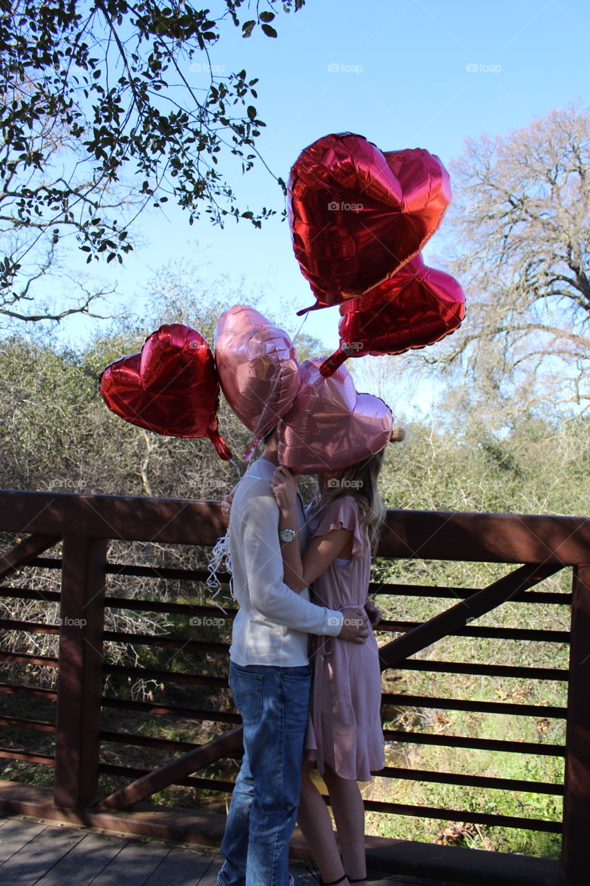 A girl and boy standing on the bridge outside in nature sharing a sweet kiss for Valentine’s Day while holding red and pink heart shaped Valentine balloons. USA, America