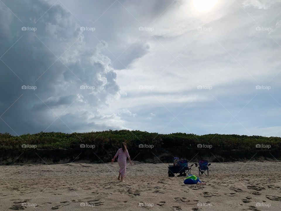 Child girl walking at the beach on the sand with multiple feet prints going towards the blue wagon and beach toys in a net bag and chairs with hill of tropical plants and grey cloudy sky on the background.