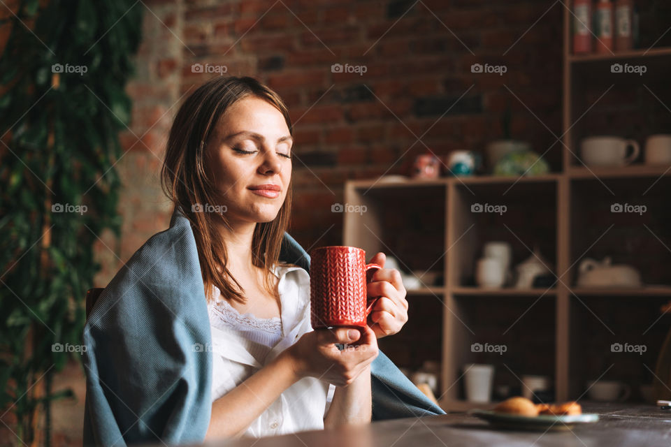 Young pretty woman in cozy grey scarf with mug of tea in hands looks out the window and rests in her studio