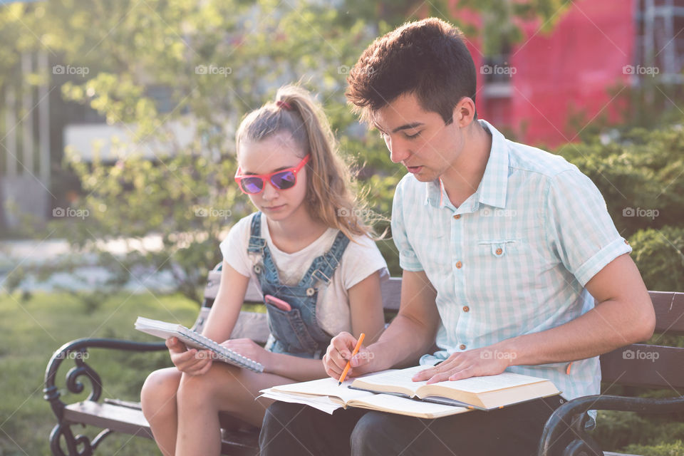 Students making the notes learning from books sitting on a bench in a park. Young boy wearing a blue shirt and dark jeans. Young blondie girl wearing jeans and sunglasses