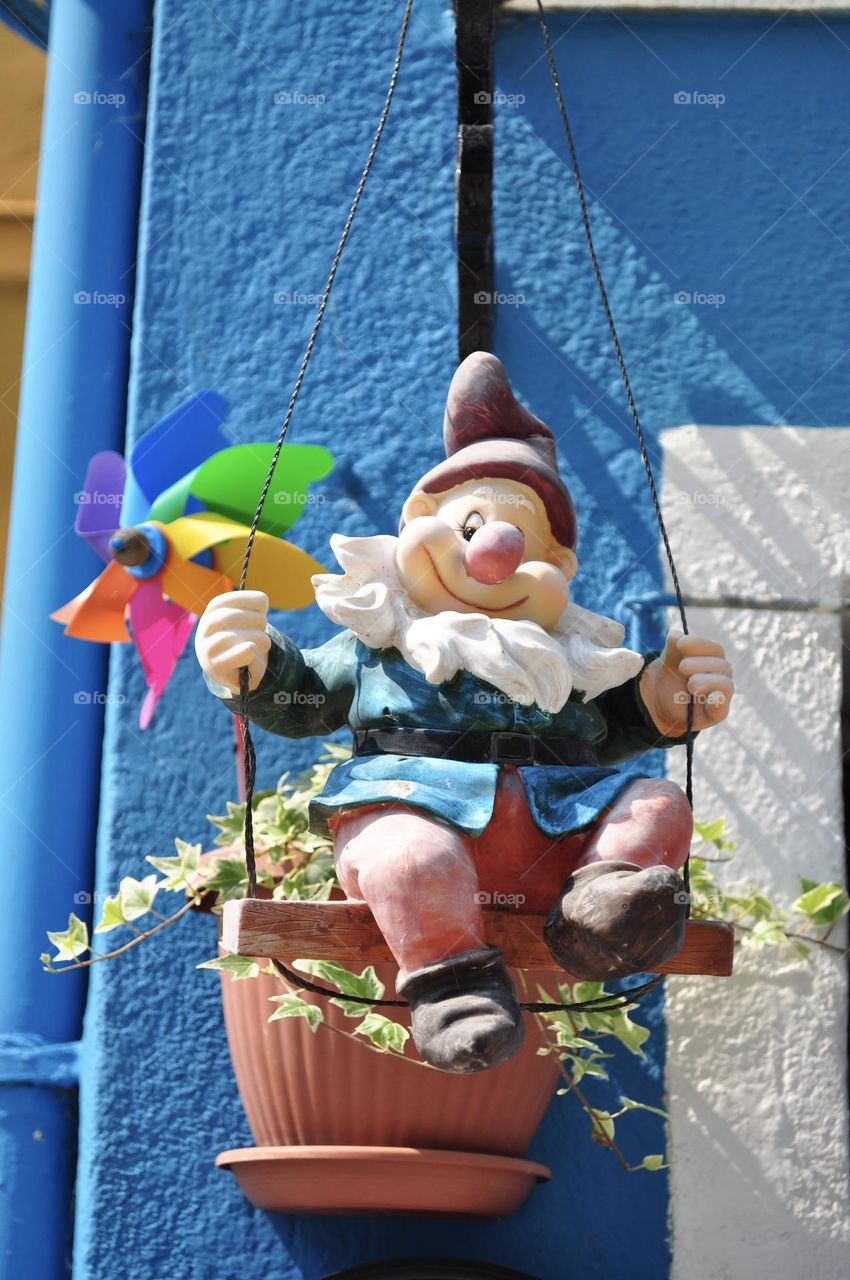 Statue of a dwarf sitting on a swing in a blue coat with a blue painted wall in the background