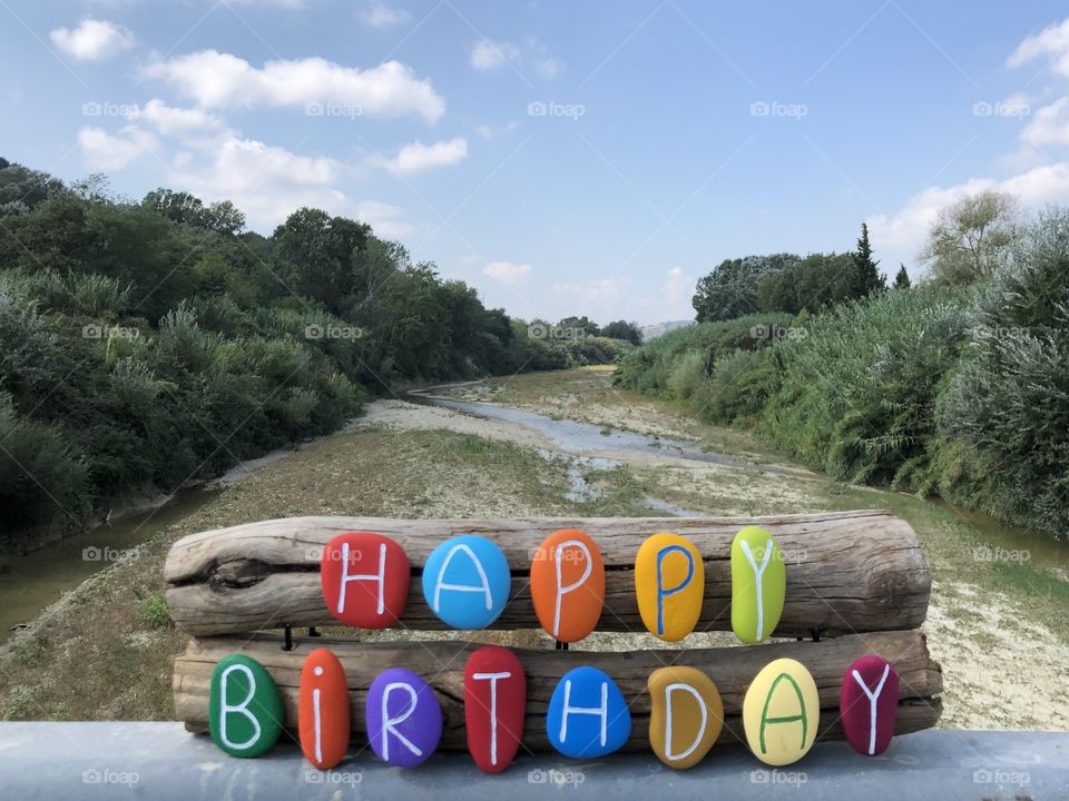 Happy Birthday message with colored stones over pieces of wood and dry river background