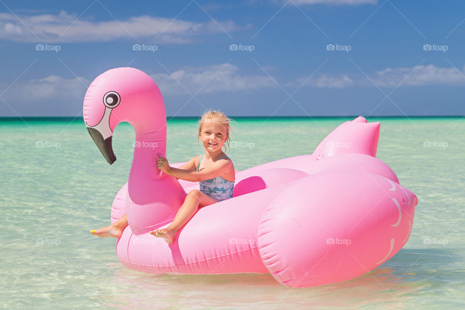 Happy little Caucasian girl with blonde hair having fun with inflatable pink flamingo on the sea