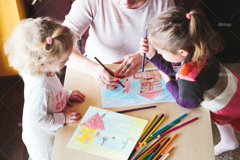 Mom with little girls drawing a colorful pictures of house and playing children using pencil crayons standing at table indoors. Shot from above