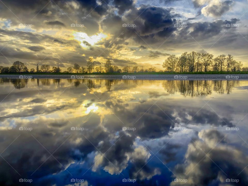Dramatic and colorful sky with white and dark clouds I between the rain during a stormy day over a Countryside landscape showing the trees in the distance, all reflected on the surface of the water in a puddle after the rain
