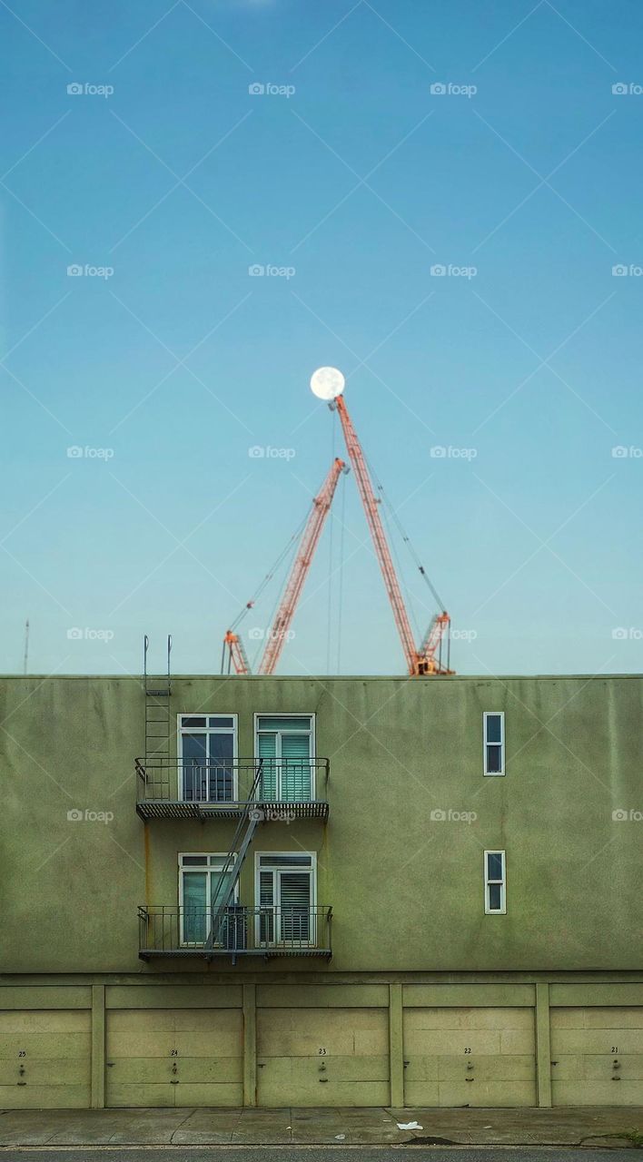 Construction cranes reaching up to a full moon behind an old building with a fire escape 