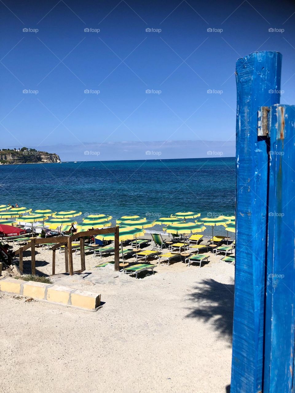 View of a beach with blue sky and sea, parasols and deckchairs installed on the sand, through a blue painted wooden gate opening onto the landscape, Tropea, Calabria, Italy