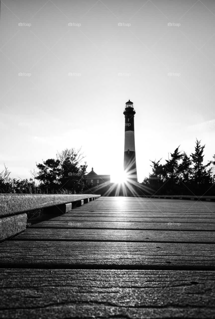 Wooden boardwalk leading to a silhouette of a tall stone lighthouse, with a light burst being created as the sun sets behind the structure. Black and white high contrast