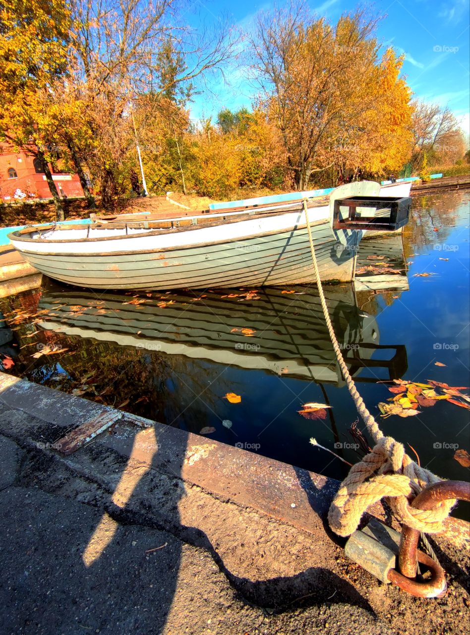Autumn.  Bank of the river.  There are yellow trees on the shore.  A wooden boat is tied to the pier with a rope.  Reflection in the water of the blue sky, boats and autumn trees