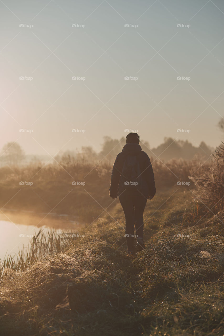 Woman walking through a meadow by a pond in the foggy morning. Sun rising above field and pond flooded with fog in the morning. Real people, authentic situations