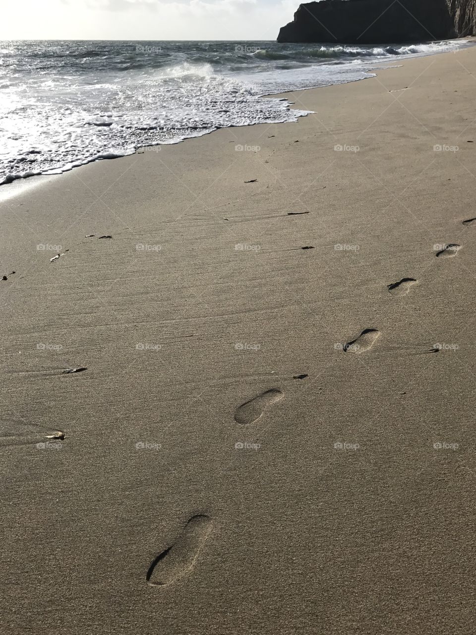 The footprints of a photographer running through damp sand on the beach, trying to catch the last of the day’s sun on the waves and cliffs of the coast. 
