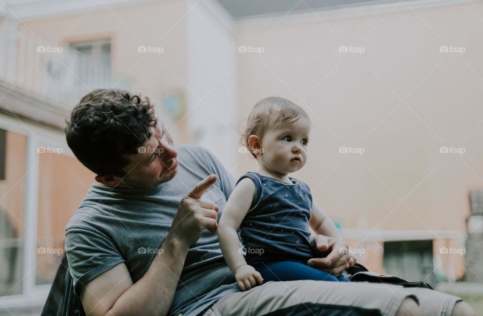 Handsome young caucasian father in work uniform with brown curly hair and a smile on his face shows with his index finger while teaching his little daughter sitting in his arms in the garden of the backyard of the house, close-up side view. Fathers