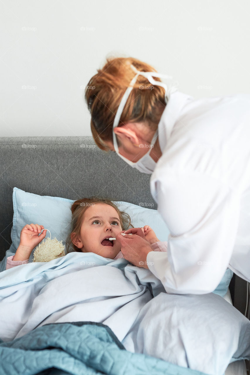 Doctor visiting little patient at home. Giving a medicine in spray to child. Woman wearing uniform and face mask. Medical treatment