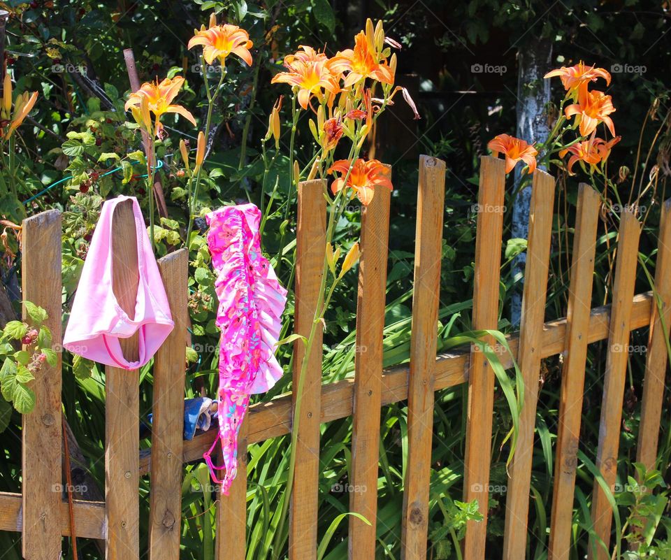 A child's  bathing suit  is drying on a wooden fence next to a garden with blooming lilies.