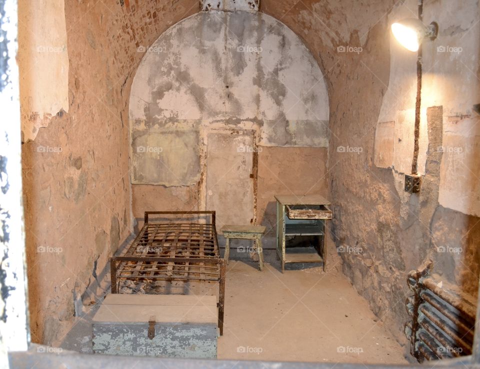 A jail cell with a metal frame bed and small dirty end table and decaying walls and a small white door under an arched ceiling 