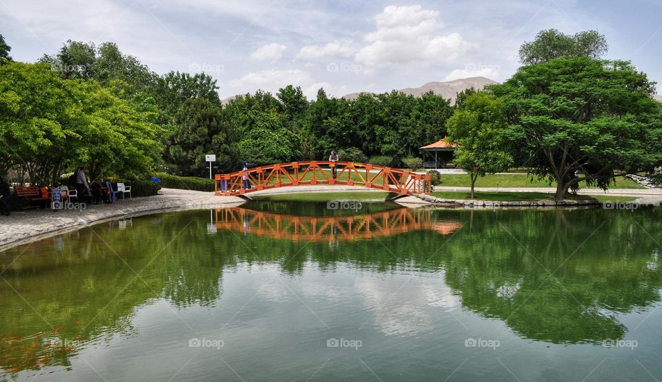 An orange bridge on the water with some trees around, and their reflection in the water.