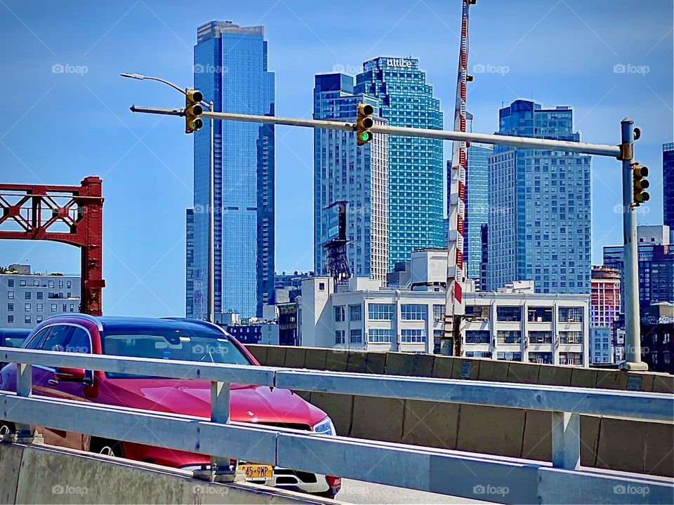 In close succession the cars are driving over the “Pulaski Bridge” that has just been reopened for traffic recognizable by the lifted red & white barrier. LIC high rises with modern glass facades on the right. 2022. Hypnotic Productions