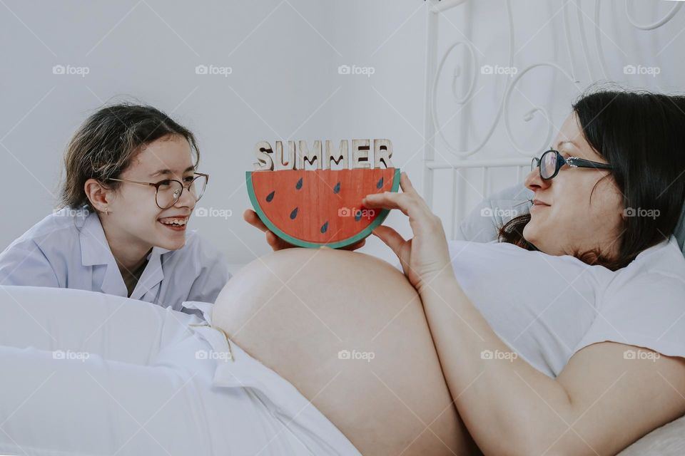 Portrait of beautiful caucasian brunette teenager girl with a happy smile in a doctor's coat sits by the bed of a pregnant mother holding a wooden slice of watermelon with the word august on her stomach, close-up side view. The concept of happy peopl