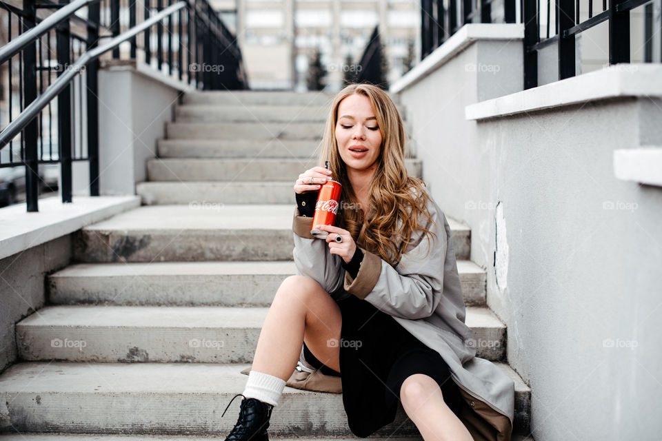 A woman in grey coat sitting on stairs and drinking Coca-Cola